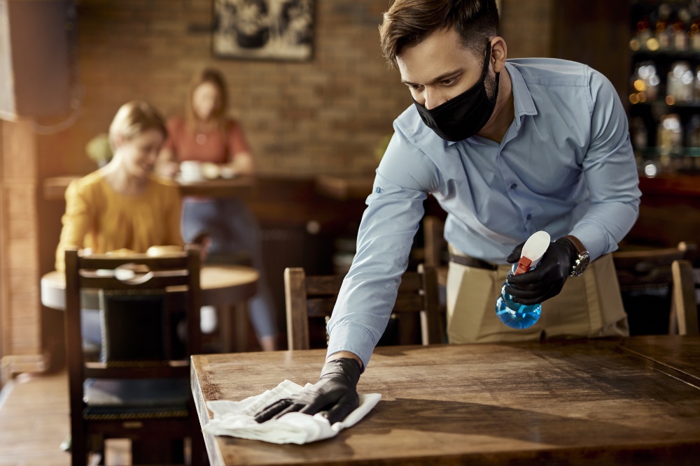 Hygiene und Gesundheitsvorsorge sind gerade im Gastronomiebereich extrem wichtig Junger Mann mit Mund-Nase-Schutz putzt Tisch in Kneipe - Eigene Bar eröffnen