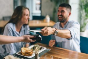 Absatz zielt auf die Menge der abgesetzten Produkte - Umsatz bezieht den Erlös mit ein Junges Paar bezahlt in einem Restaurant mit Handy - so entsteht Umsatz