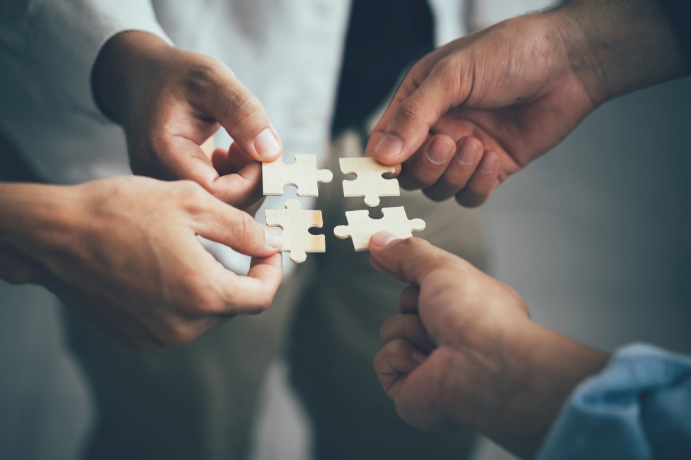 4 Menschen halten jeweils ein Puzzleteil in der Hand - Symbol für eine Kooperation - Welche Vorteile bietet ein Joint Venture?